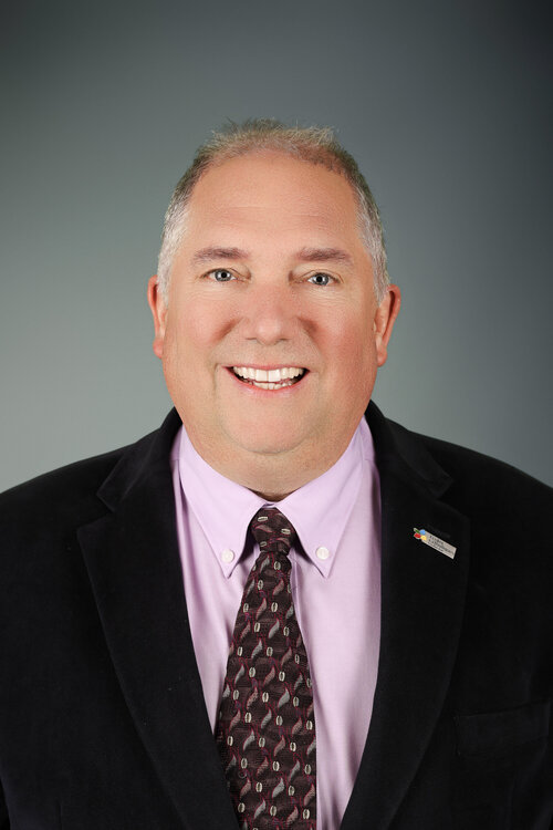 Portrait professionnel de Robert Rainboth souriant, vêtu d’un veston à carreaux mauves, d’une chemise bleue et d’une cravate violette.