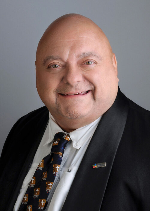 Portrait professionnel de Denis Forget souriant, vêtu d’un veston noir, chemise blanche et cravate ornée de croix dorées.