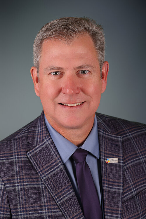 Portrait professionnel de Robert Rainboth souriant, vêtu d’un veston à carreaux mauves, d’une chemise bleue et d’une cravate violette.
