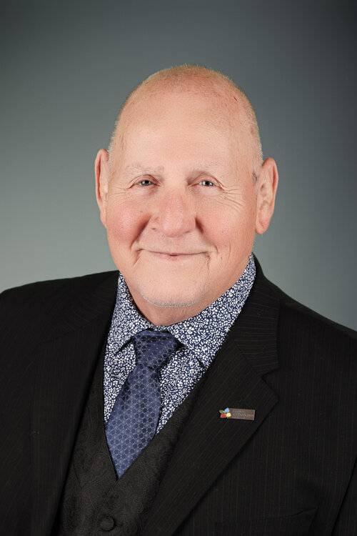 Portrait professionnel d’André Thibodeau souriant, vêtu d’un complet noir, d’une chemise à motifs floraux et d’une cravate bleue.
