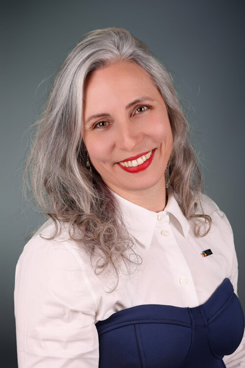 Julie Cardinal souriante, vêtue d’un chemisier blanc et corsage bleu, sur fond dégradé neutre.