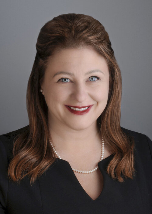 Marie-France Paquette souriante, vêtue de noir avec un collier de perles, devant un fond gris neutre.