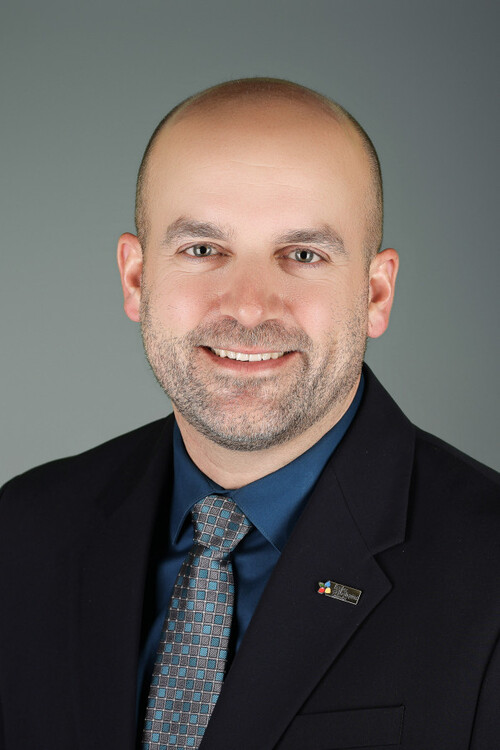  Portrait professionnel de Marc Bertrand souriant, vêtu d’un veston noir, chemise bleue foncée et cravate à motifs gris.