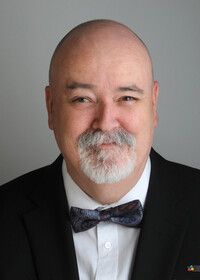 ortrait professionnel de Claude Lalonde souriant, vêtu d’un veston foncé, d’une chemise blanche et d’un nœud papillon à motifs, sur un fond uni gris.