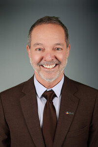 Portrait professionnel de Michel Charron souriant, vêtu d’un complet brun et d’une chemise pâle avec cravate assortie.