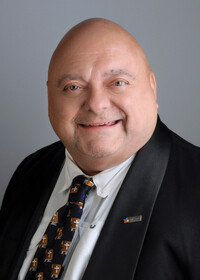 Portrait professionnel de Denis Forget souriant, vêtu d’un veston noir, chemise blanche et cravate ornée de croix dorées.