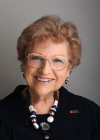 Portrait professionnel de Monique Briand souriante, portant des lunettes dorées, un collier noir et blanc et un haut foncé.