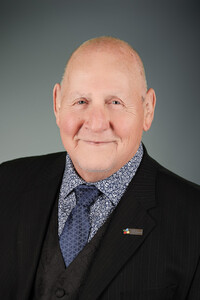 Portrait professionnel d’André Thibodeau souriant, vêtu d’un complet noir, d’une chemise à motifs floraux et d’une cravate bleue.