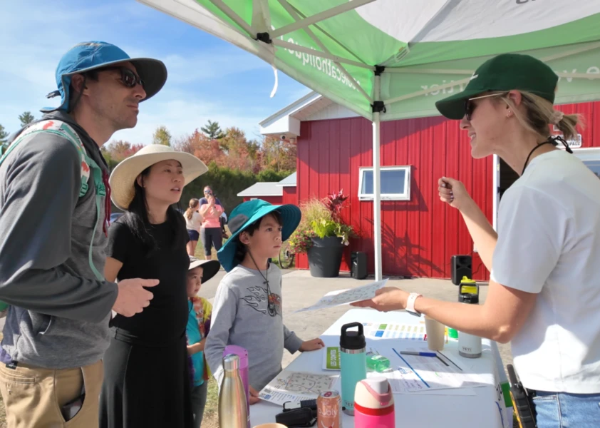 Une animatrice présente une activité à une famille sous un chapiteau, devant un bâtiment rouge, lors d’un événement extérieur ensoleillé.