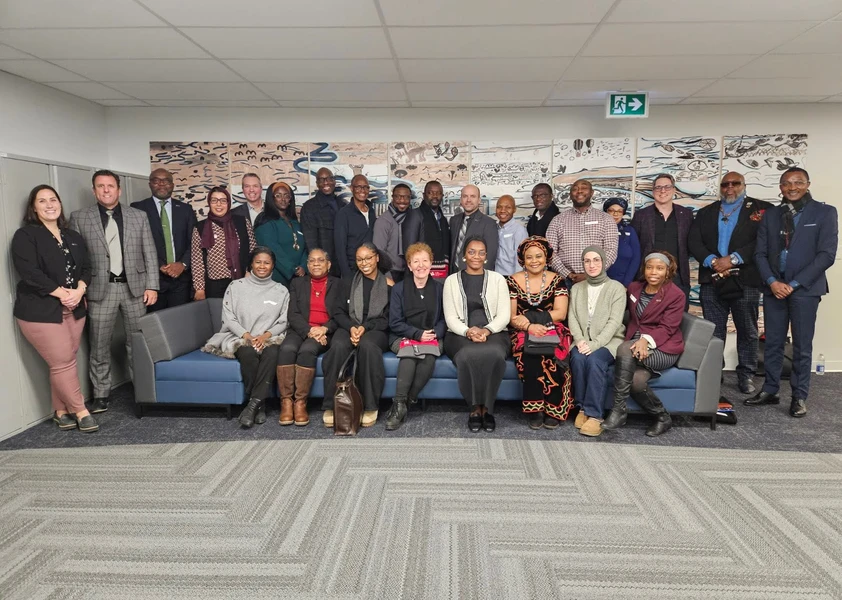 Photo de groupe d’une vingtaine d’adultes issus de la diversité, certains assis sur un divan et d’autres debout derrière, réunis à l’intérieur et souriant face à la caméra.