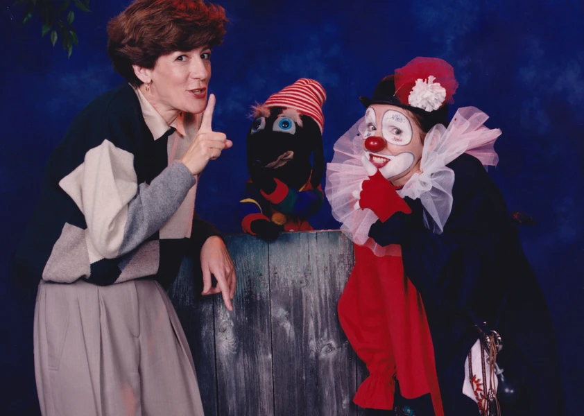 Suzanne Pinel fait un geste de silence à côté d’une marionnette et d’un clown souriant, sur un fond de studio bleu.