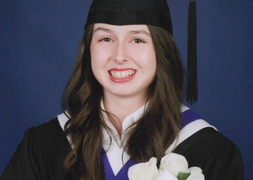 Portrait officiel de fin d’études de Kaylee Gagné portant une toge noire et un mortier de diplômée. Elle tient un bouquet de roses blanches accompagné d’un rouleau de diplôme marqué « 2025-2026 » devant un fond bleu uni et sourit à la caméra.