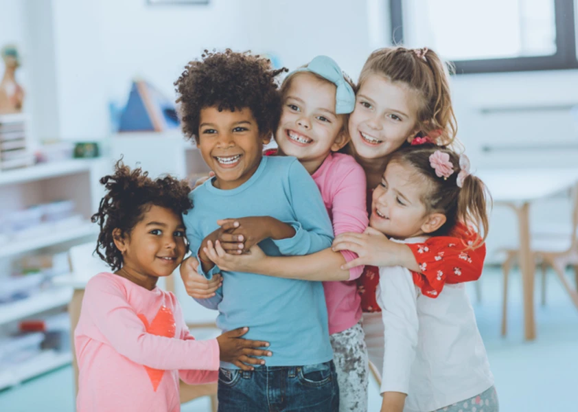Cinq jeunes enfants souriants se font un câlin dans une salle de classe.