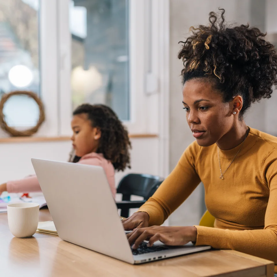 Parent travaillant sur un ordinateur portable à une table, avec un élève à l’arrière-plan en train de dessiner avec des crayons de couleur.