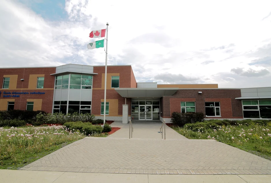 Façade de l’École élémentaire catholique Saint-Rémi, bâtiment moderne en briques rouges et panneaux gris avec de grandes fenêtres. Un parterre fleuri et deux drapeaux, canadien et franco-ontarien, ornent l’entrée principale.