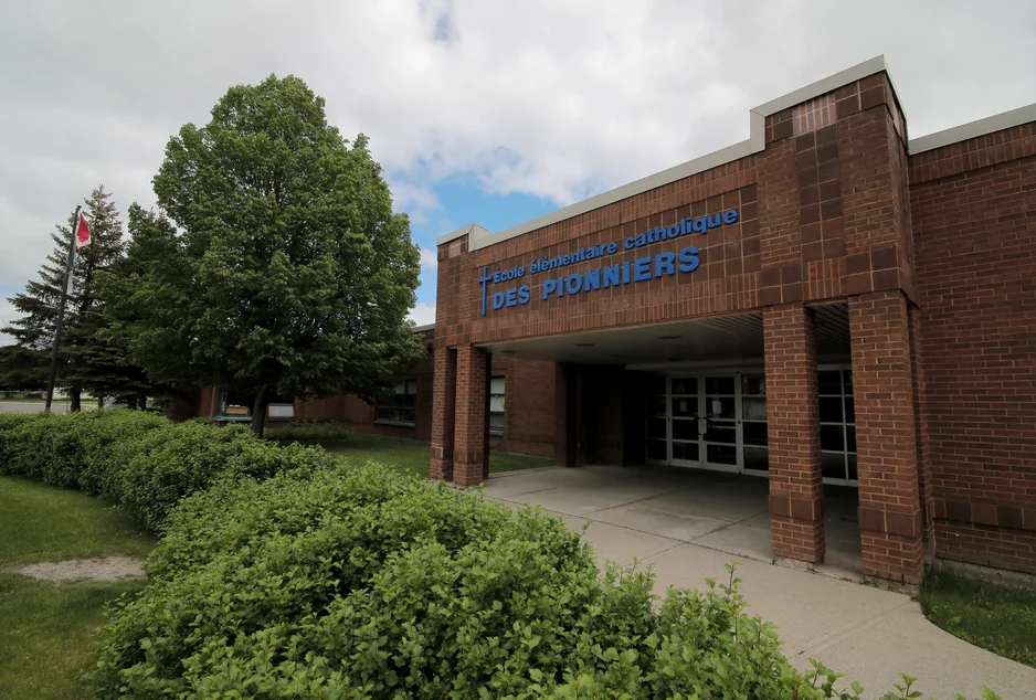 açade de l’École élémentaire catholique des Pionniers, bâtiment en briques rouges avec le nom de l’école en lettres bleues. Des arbustes et un grand arbre bordent l’entrée. Un drapeau du Canada flotte à gauche.