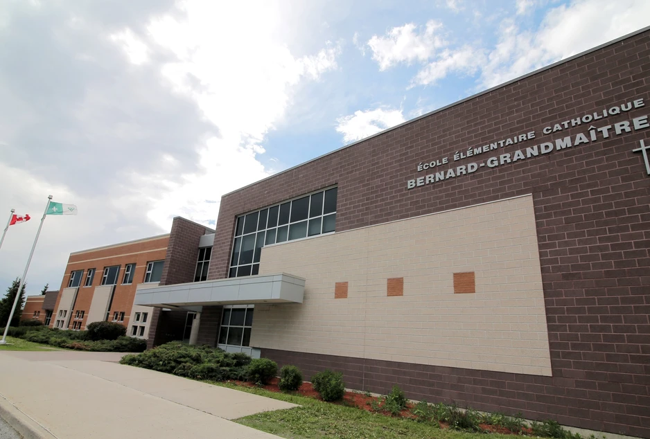 Façade de l’École élémentaire catholique Bernard-Grandmaître, bâtiment moderne en briques brunes et beiges avec de grandes fenêtres. Trois drapeaux flottent devant l’école : celui de l’Ontario, du Canada et de la francophonie. Un trottoir borde l’avant de l’école, avec des arbustes paysagers le long de la façade.