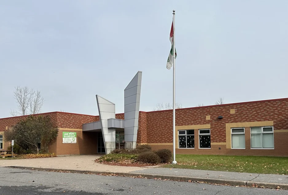 Façade de l’École élémentaire catholique L’Étoile-de-l’Est, bâtiment en briques rouges avec une entrée moderne aux structures métalliques argentées. Deux drapeaux flottent devant l’école.