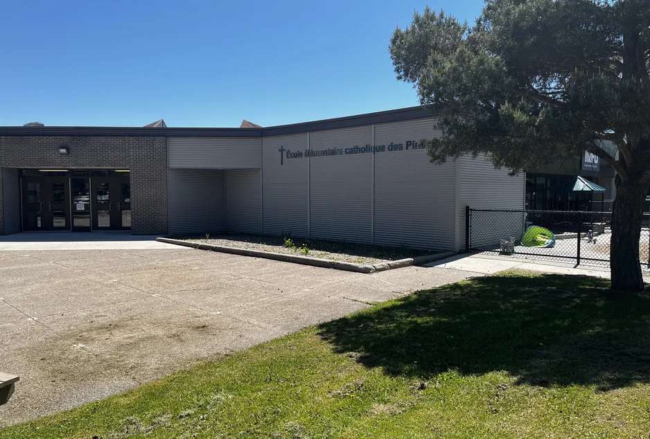  Façade de l’École élémentaire catholique des Pins, bâtiment à revêtement métallique gris et briques foncées, avec une entrée vitrée à gauche. Une cour clôturée avec du mobilier extérieur est visible à droite, partiellement ombragée par un grand arbre.