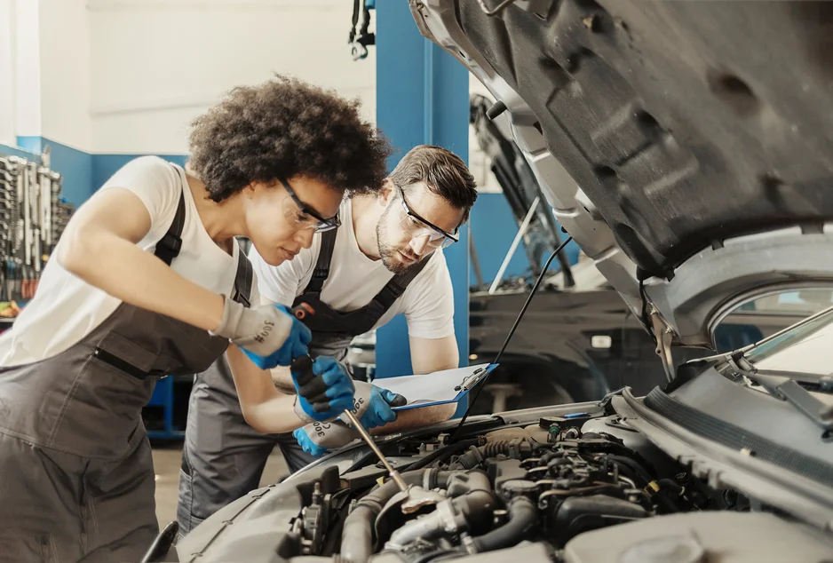 Un élève en tenue de travail utilise un tournevis pour intervenir sous le capot ouvert d’une voiture, pendant qu’un autre élève observe attentivement en tenant une tablette ou une planchette. Les deux portent des lunettes de protection et des gants, dans un atelier mécanique bien équipé. L’image illustre un apprentissage pratique en mécanique automobile.