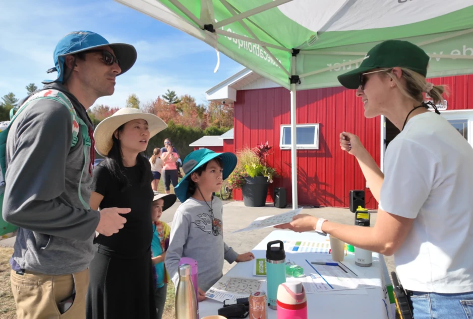 Une animatrice présente une activité à une famille sous un chapiteau, devant un bâtiment rouge, lors d’un événement extérieur ensoleillé.