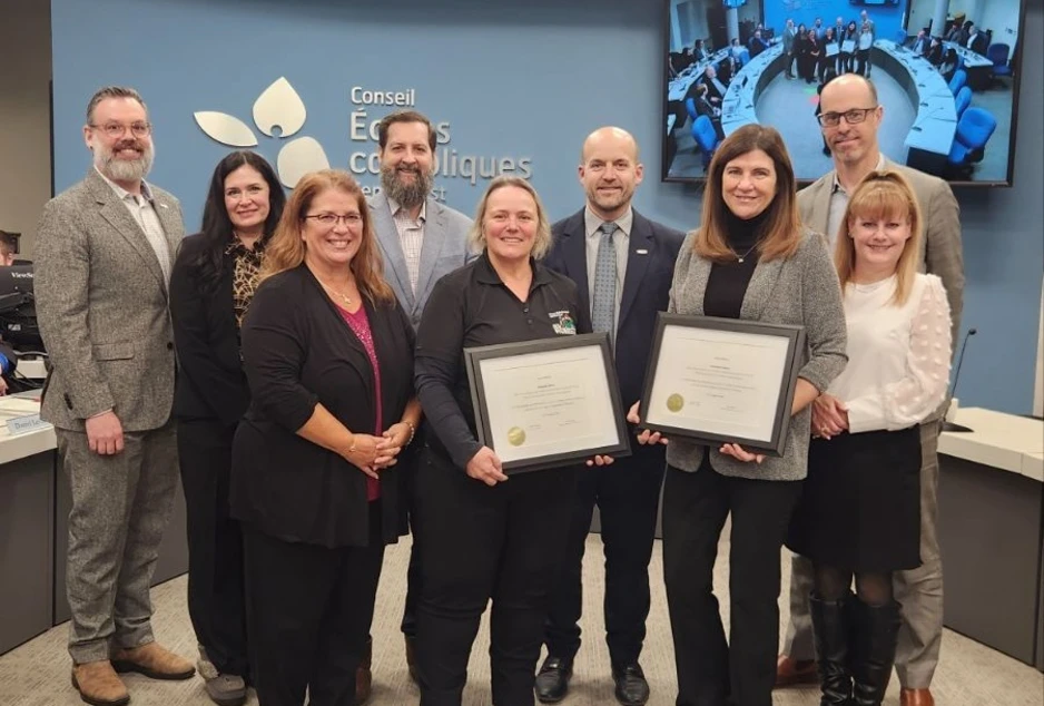 Groupe de huit adultes posant devant le mur du Conseil des écoles catholiques, deux femmes tenant des certificats encadrés lors d’une remise officielle.