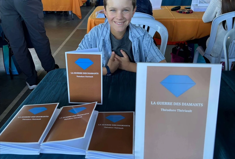 Jeune élève souriant assis derrière une table présentant plusieurs exemplaires de son livre La guerre des diamants lors d’un salon ou d’une activité littéraire scolaire.