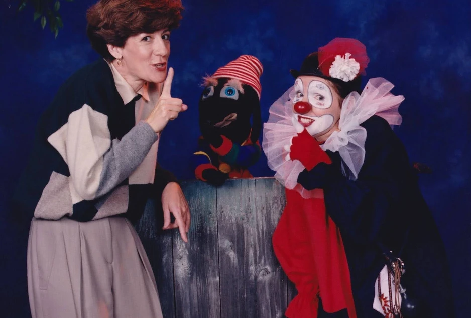 Une femme fait un geste de silence avec le doigt sur les lèvres, un personnage marionnette coloré apparaît au centre, et un clown maquillé en rouge, blanc et noir sourit en imitant le même geste.