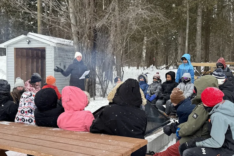 Groupe d’élèves habillés en tenue d’hiver écoutant leur enseignante qui anime une activité extérieure près d’un feu, en forêt enneigée.
