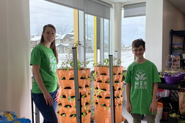 Élève et enseignante souriants devant des tours de culture orange remplies de jeunes plantes, dans une classe lumineuse.