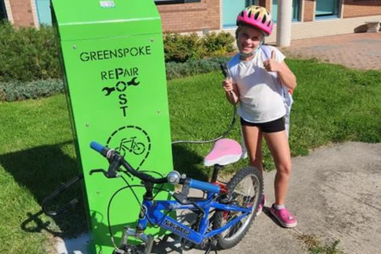 Jeune élève souriante avec son vélo devant une station verte de réparation installée à l’entrée de l’école.