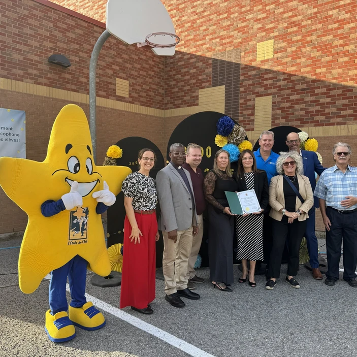 Groupe d’adultes posant à l’extérieur d’une école aux côtés d’une mascotte en forme d’étoile, lors d’une célébration.
