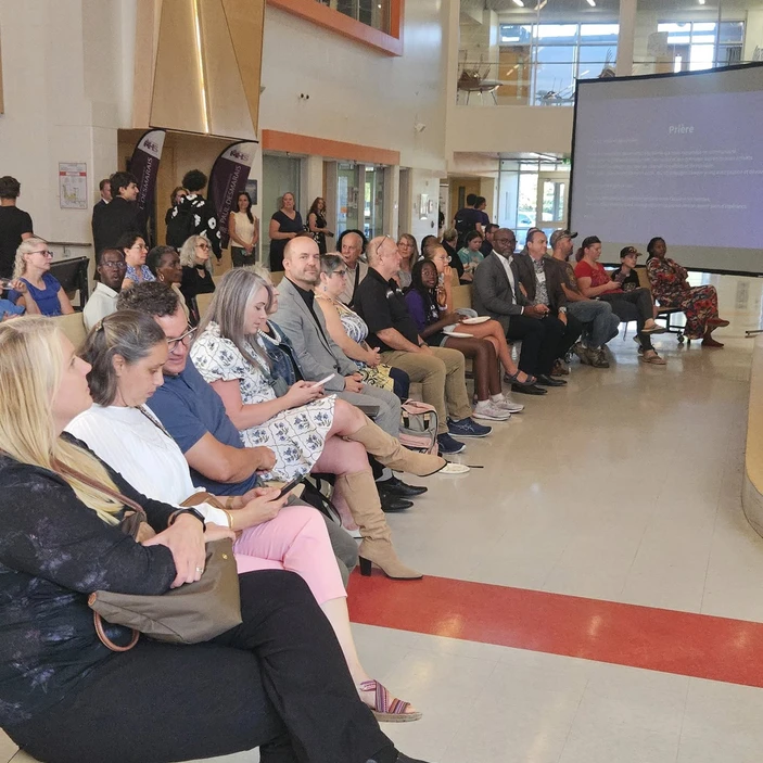 Public assis dans le hall lumineux d’une école, assistant à une présentation projetée sur écran.