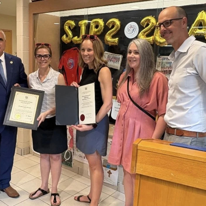 Deux personnes tiennent des certificats de reconnaissance devant un décor festif composé de ballons dorés marquant les 20 ans de l’école, d’articles souvenirs et de photos historiques. L’ambiance est chaleureuse et souligne les contributions de la communauté scolaire à cet anniversaire.