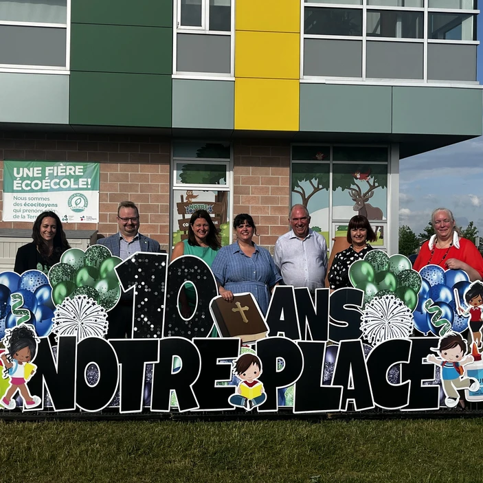Photo de groupe prise devant l’École élémentaire catholique Notre-Place à l’occasion de son 10e anniversaire. Sept adultes souriants posent derrière une grande enseigne décorative portant l’inscription « 10 ans Notre-Place », entourée d’illustrations d’élèves, de livres, de matériel scolaire et de ballons festifs.