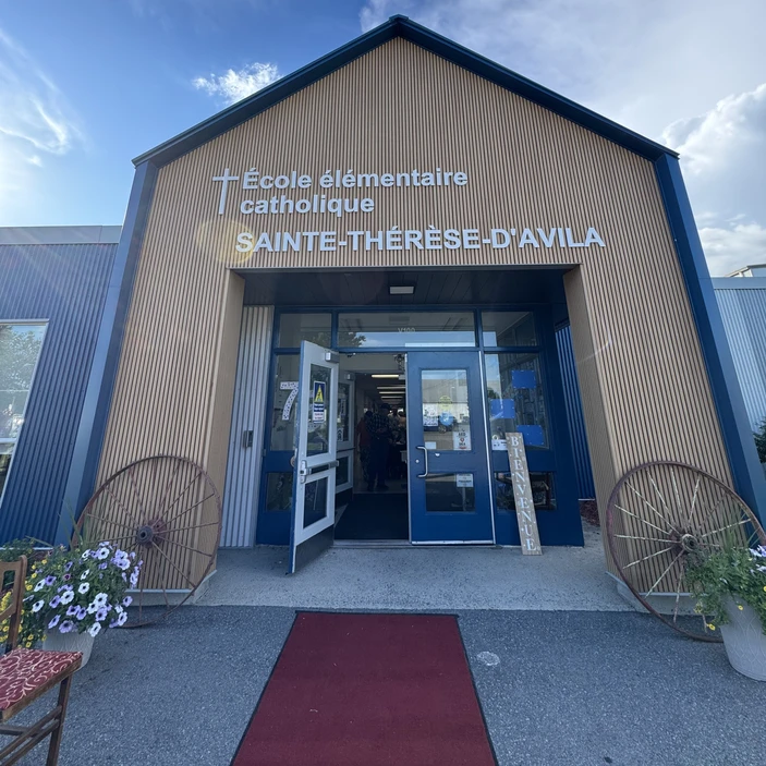 Vue de l'entrée principale de l'École élémentaire catholique Sainte-Thérèse-d'Avila. La façade est revêtue de lattes de bois verticales avec le nom de l'école affiché en grandes lettres blanches. Un tapis rouge mène aux portes d'entrée ouvertes, encadrées de deux grandes roues de chariot décoratives et de bacs de fleurs.