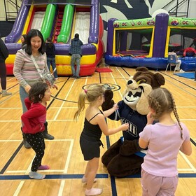 Enfants dans un gymnase autour d’une mascotte, avec des jeux gonflables en arrière-plan.