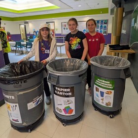 Trois élèves posent devant des bacs de tri pour les déchets, le papier-carton et les matières organiques dans leur école.